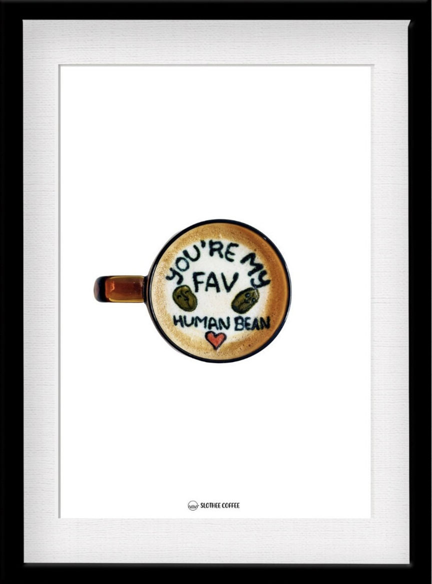 latte art print by jess lui in a frame. latte art has written text on it "you're my fav human bean" with two beans colored brown and a red heart. art drawn (or etched) on white milk foam with a border of coffee around it. mug handle on left side.